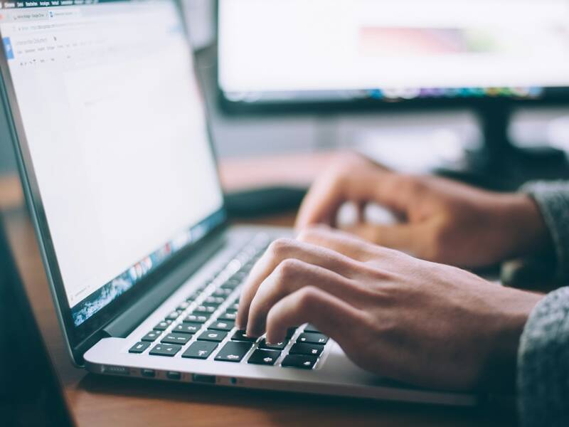 Eine Person tippt auf der Tastatur eines Laptops, während im Hintergrund unscharf ein weiterer Bildschirm zu sehen ist.