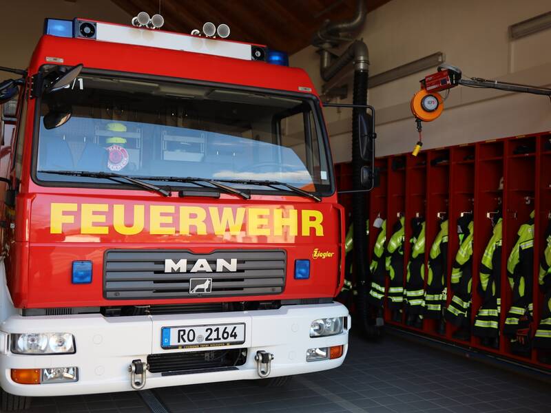 Ein rotes Feuerwehrfahrzeug steht in einer Fahrzeughalle neben aufgereihten Einsatzjacken der Feuerwehr.