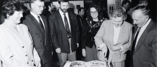 Sechs lächelnde Personen, darunter Altbürgermeister Dr. Keitz, stehen um einen Tisch herum, auf dem eine große, rechteckige Torte mit einem Bild und Schriftzug obenauf liegt, während eine Person ein Messer zum Anschneiden der Torte hält.