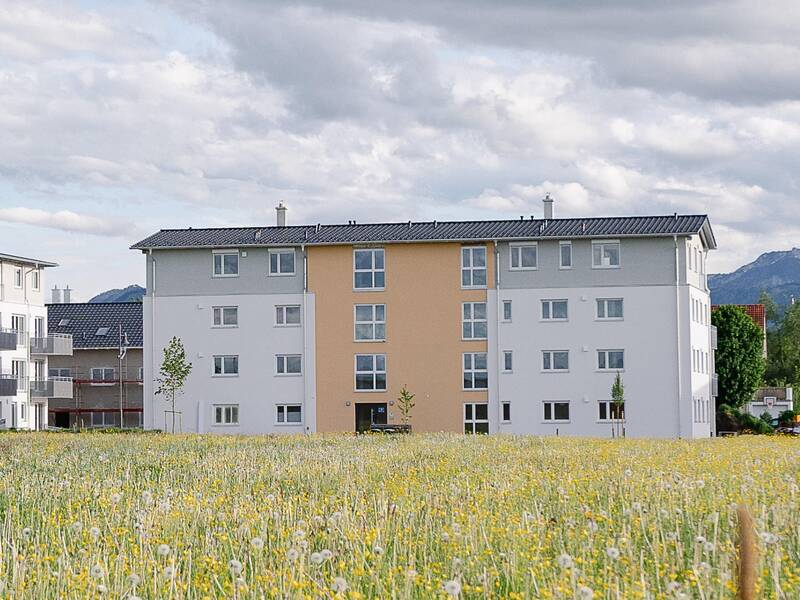 Ein mehrstöckiges Wohngebäude steht auf einem Feld mit blühenden Wildblumen, während im Hintergrund Berggipfel zu sehen sind.