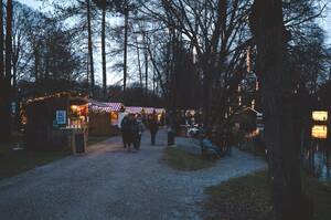 Das Bild zeigt einen festlich beleuchteten Weihnachtsmarkt in einem Park bei Dämmerung, mit Menschen, die entlang von Verkaufsbuden spazieren.